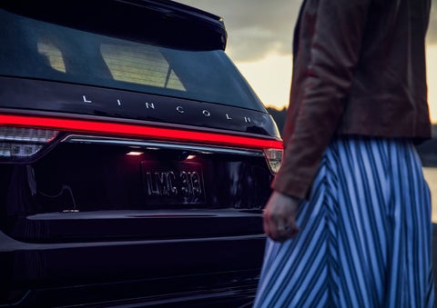 A person is shown near the rear of a 2024 Lincoln Aviator® SUV as the Lincoln Embrace illuminates the rear lights | Lincoln Demo 3 in Wooster OH